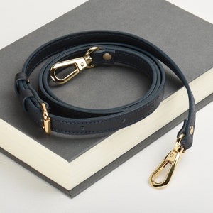 Peut inclure: Une sangle en cuir bleu foncé avec des ferrures dorées. La sangle est enroulée et posée sur un livre gris.