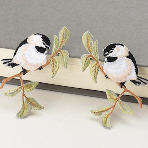 May include: Two embroidered patches featuring birds perched on branches with leaves. The birds have black and white markings with tan bellies. The branches are brown, and the leaves are green and yellow. The patches are likely for clothing or craft projects.