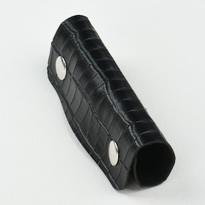 May include: Black leather wrist cuff with a crocodile texture. The cuff is secured with two silver-colored snap closures. The cuff is curved and appears to be designed to wrap around the wrist.