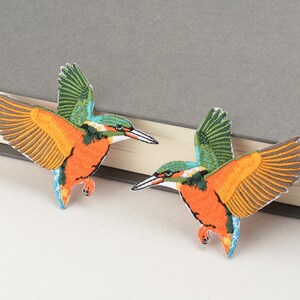 Puede incluir: Dos parches bordados de un martín pescador en vuelo. Las aves tienen cuerpos naranjas, alas verdes y detalles azules. Los parches están detallados con intrincadas puntadas y se colocan sobre un fondo neutro.