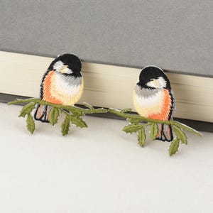 Op de afbeelding: Twee geborduurde vogelpatches. Elke vogel heeft een zwarte kop, een witte en oranje borst en grijze en zwarte vleugels. Ze zitten op een groene geborduurde tak met bladeren. De patches liggen op een wit oppervlak.