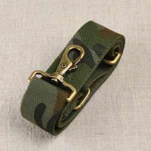 Puede incluir: Correa de lona con estampado de camuflaje con herrajes metálicos de color latón antiguo. La correa presenta un diseño de camuflaje verde, marrón y negro. Los herrajes metálicos incluyen un cierre giratorio y dos bucles rectangulares.
