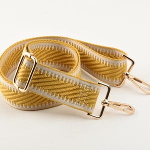 May include: A yellow and white patterned bag strap with gold-colored metal hardware. The strap features a woven chevron design in yellow and white. The metal hardware includes rectangular rings and swivel clasps.