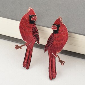 Peut inclure: Deux écussons brodés représentant des oiseaux cardinaux rouges. Les oiseaux ont des accents noirs, des pattes marron et une bordure dorée. Ils sont perchés sur une branche. Les écussons sont sur une surface blanche avec un livre gris en arrière-plan.