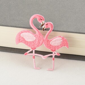 Pode incluir: Patch bordado com dois flamingos cor-de-rosa num abraço amoroso. Os flamingos têm detalhes em rosa claro nas asas e bicos castanhos. O patch é costurado com um padrão de ponto detalhado, perfeito para adicionar um toque tropical a roupas ou acessórios.