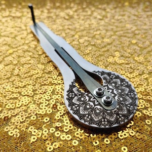 May include: A silver-colored, hand-held musical instrument with a mandala design etched on the soundboard. The instrument is resting on a gold sparkly surface.