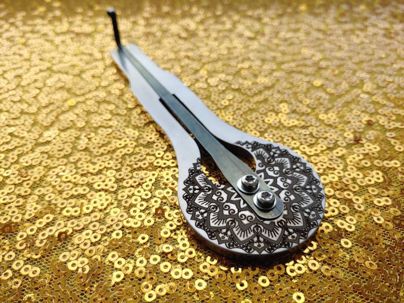 May include: A silver-colored, hand-held musical instrument with a mandala design etched on the soundboard. The instrument is resting on a gold sparkly surface.