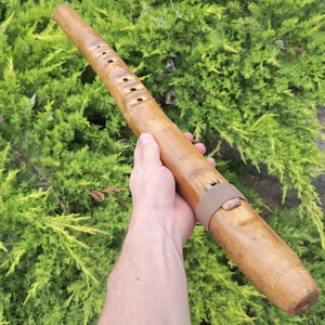 Puede incluir: Una flauta de madera hecha a mano con múltiples agujeros para los dedos y una correa de cuero. El instrumento es de color marrón claro y parece estar hecho de una sola pieza de madera. La flauta se sostiene contra un fondo de follaje verde.