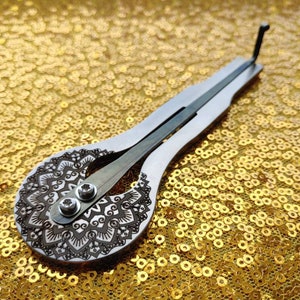 May include: A silver and wood musical instrument with a mandala design on the soundboard. The instrument is lying on a gold sequined surface.