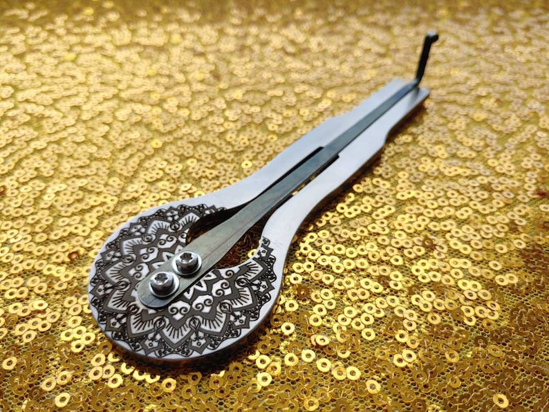 May include: A silver and wood musical instrument with a mandala design on the soundboard. The instrument is lying on a gold sequined surface.