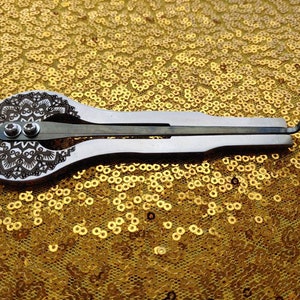 May include: A silver-colored, hand-crafted musical instrument called a  'thumb piano' or 'kalimba' with a decorative mandala design on the soundboard. The instrument has a single row of tines and a small handle.