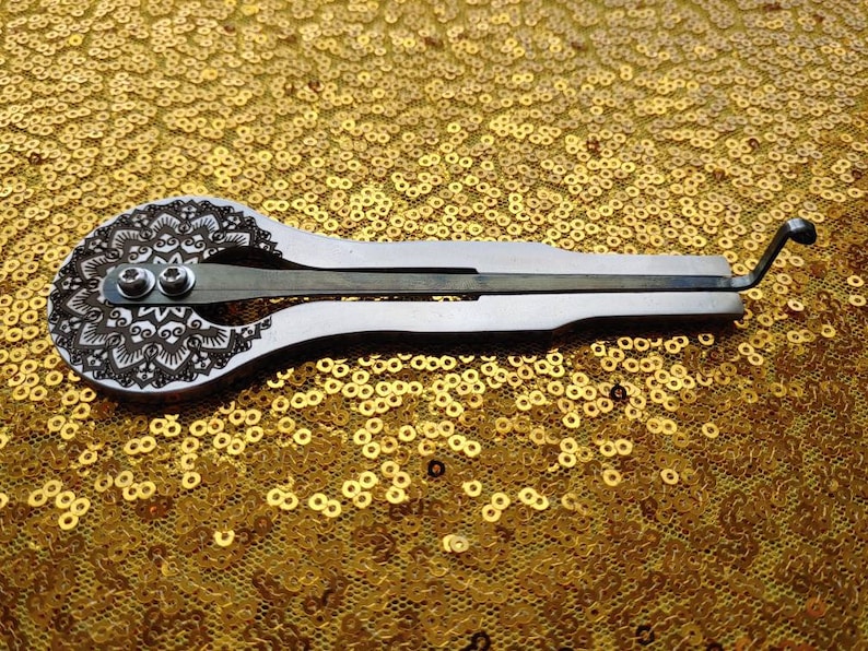 May include: A silver-colored, hand-crafted musical instrument called a  'thumb piano' or 'kalimba' with a decorative mandala design on the soundboard. The instrument has a single row of tines and a small handle.