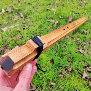 Puede incluir: Flauta de madera hecha a mano con acabado de madera natural. La flauta tiene un cuerpo rectangular con agujeros para los dedos y una banda negra alrededor de la boquilla. Se adjunta un cordón negro. El instrumento se sostiene sobre un fondo de hierba verde.