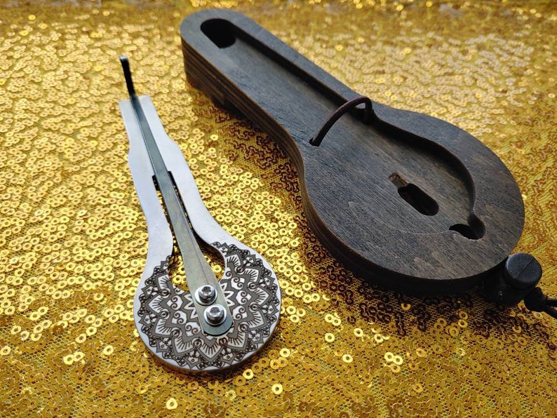 May include: A silver-colored,  hand-crafted,  musical instrument called a  'thumb piano' or 'kalimba' with a decorative mandala design. The instrument is resting on a gold sequined surface.