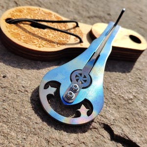 Könnte beinhalten: Ein blau-silbernes Musikinstrument, eine Maultrommel, liegt auf einem Holzkoffer mit dekorativem Design. Die Maultrommel hat ein Stern- und Monddesign. Das Instrument ist etwa 10 cm lang.