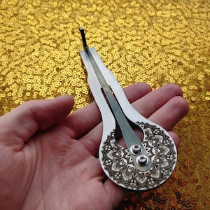 May include: A silver-colored, hand-held musical instrument with a mandala design. The instrument has two strings and a small hole at the top.