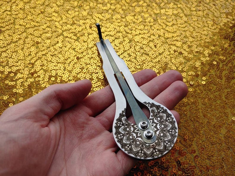 May include: A silver-colored, hand-held musical instrument with a mandala design. The instrument has two strings and a small hole at the top.