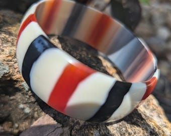 May include: A colorful, striped bangle bracelet. The bracelet features alternating bands of red, white, black, and gray. The chunky bracelet is made of a glossy material and is displayed on a textured rock surface.