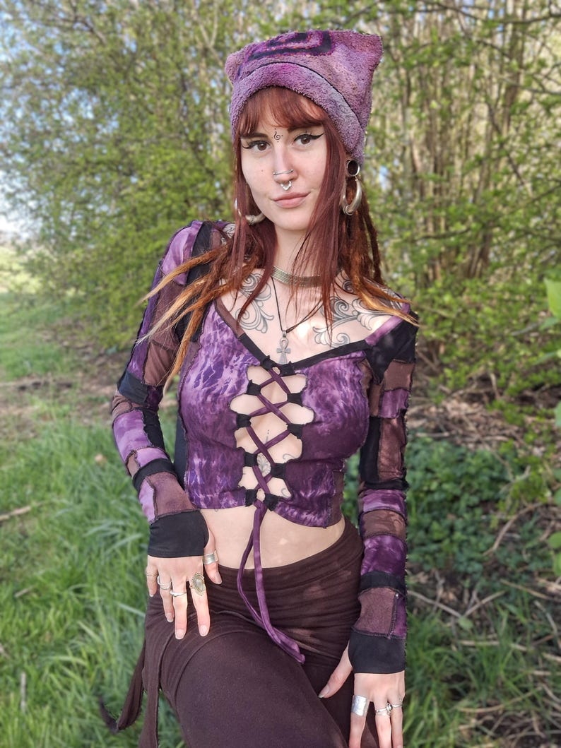 May include: A purple tie-dye crop top with long sleeves and a lace-up front. The top is paired with brown trousers and a matching purple hat. The outfit has a bohemian, alternative style, with patchwork details and a unique aesthetic.