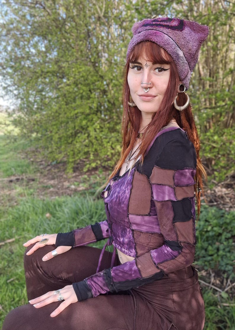 May include: A patchwork top in shades of purple, brown, and black, with matching brown trousers. The outfit is completed by a purple beanie. The style is bohemian and unique.