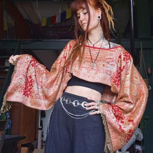 Op de afbeelding: Een cropped, rood en goudkleurig patroon top met lange, vloeiende mouwen en een gefranjerde zoom. De top wordt gecombineerd met een zwarte broek en een zilveren kettingriem. De outfit heeft een bohemien esthetiek.