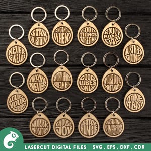 May include: A collection of wooden keychains with silver key rings. Each keychain is teardrop-shaped and engraved with motivational phrases like "Stay Positive," "Never Give Up," and "Good Vibes Only." The keychains are arranged on a dark wooden surface.