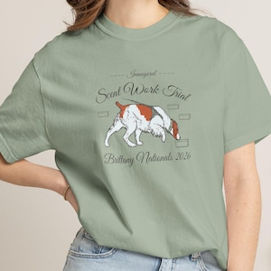 Könnte beinhalten: Ein salbeigrünes Kurzarm-T-Shirt mit einer Grafik eines weiß-braunen Brittany-Hundes, der am Boden schnüffelt. Der Text lautet Inaugural Scent Work Trial Brittany Nationals 2026, umgeben von kleinen rechteckigen Kästchen um die Hundeabbildung.