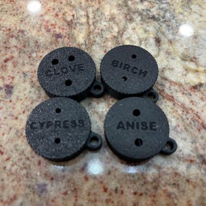 May include: Four black round disc-shaped aromatherapy diffusers with holes and a small loop on the side. The diffusers are labelled "CLOVE", "BIRCH", "CYPRESS", and "ANISE".