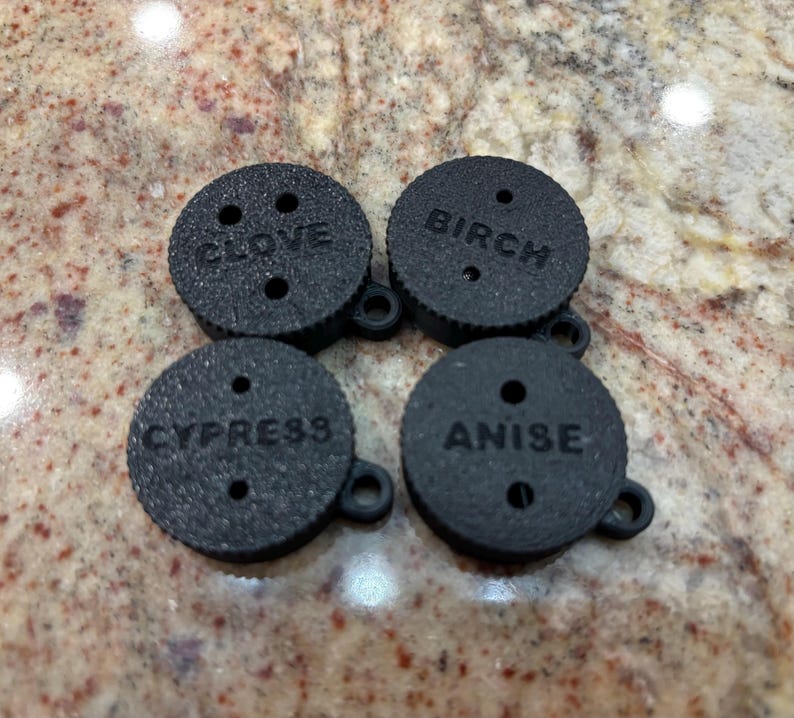 May include: Four black round disc-shaped aromatherapy diffusers with holes and a small loop on the side. The diffusers are labelled "CLOVE", "BIRCH", "CYPRESS", and "ANISE".