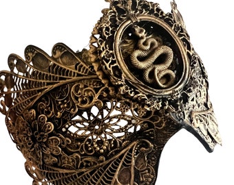 May include: A gold-colored metal mask with intricate floral designs and a snake in a circular frame. The mask is made of delicate, openwork metal.