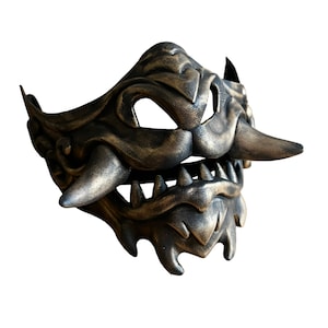 May include: A black and gold mask with a fierce expression. The mask has sharp teeth, a long nose, and a textured surface.