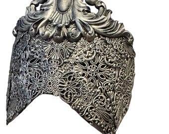 May include: A silver-colored, ornate headpiece with intricate floral and scrollwork designs. The headpiece features a central oval embellishment and a detailed, openwork pattern throughout, creating a vintage or fantasy aesthetic.