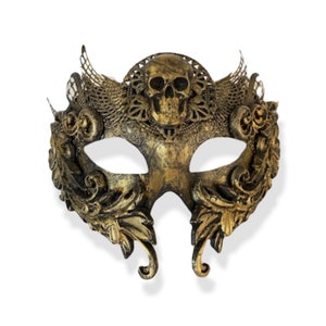 Op de afbeelding: Een goudkleurig masker voor een gemaskerd bal met een schedelontwerp in het midden bovenaan. Het masker heeft sierlijke details, waaronder bloemen- en filigraanpatronen. De ooggaten zijn ovaal van vorm en het masker heeft een gebogen vorm om op het gezicht te passen.