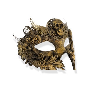 Op de afbeelding: Een goudkleurig masker met een schedelontwerp. Het masker heeft sierlijke details, waaronder bloem- en vleugelachtige accenten. Het masker is gemaakt van een metallic materiaal en is ontworpen om de ogen en neus te bedekken.