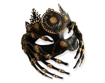 May include: Black masquerade mask with gold accents and a skeletal hand design. The mask has a crown-like top and intricate details.