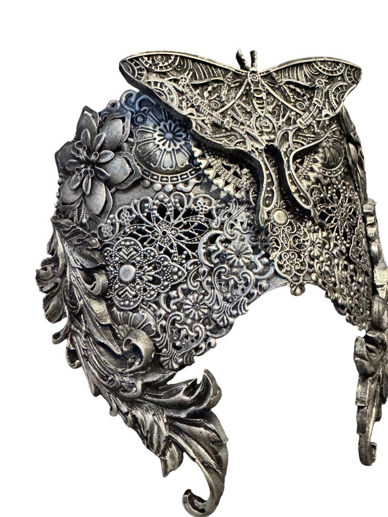 May include: Silver-toned headpiece with intricate floral, leaf, and lace-like patterns. A detailed moth adorns the top center. The headpiece has a curved shape and is likely made of metal.