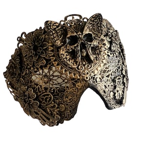 Puede incluir: Una m&aacute;scara de carnaval dorada con un dise&ntilde;o de calavera y mariposa. La m&aacute;scara est&aacute; intrincadamente detallada con patrones florales y tiene un respaldo de terciopelo negro.