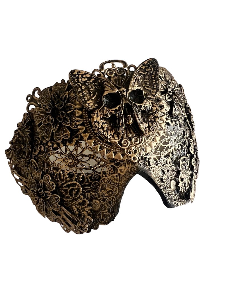Puede incluir: Una m&aacute;scara de carnaval dorada con un dise&ntilde;o de calavera y mariposa. La m&aacute;scara est&aacute; intrincadamente detallada con patrones florales y tiene un respaldo de terciopelo negro.
