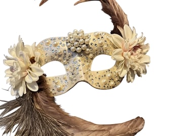 May include: An ivory-colored masquerade mask adorned with faux pearls and rhinestones. The mask features two cream-colored fabric flowers and is embellished with brown feathers. A cluster of pearls sits above the nose bridge.
