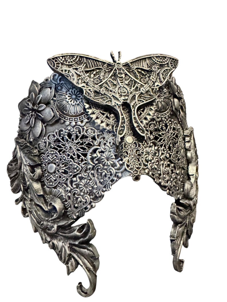 May include: A silver-toned headpiece with intricate floral, leaf, and lace-like patterns. A detailed butterfly design is centered on the crown. The headpiece has a gothic or fantasy aesthetic.
