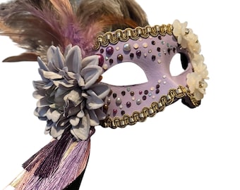 May include: A lavender masquerade mask with gold trim, adorned with pearls, rhinestones, and a large gray flower. The mask features a tassel and is embellished with feathers in shades of gray, pink, and purple.