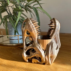 May include: A gold-coloured dinosaur skull planter with a wooden insert. The planter is made of 3D printed plastic and has a detailed design.