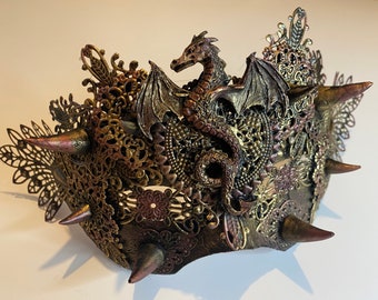 May include: A gold and brown metal mask with a dragon design. The mask is decorated with intricate details and has sharp spikes around the edges.
