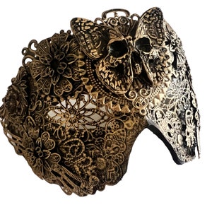 Puede incluir: Una m&aacute;scara de metal dorado con un dise&ntilde;o de calavera y mariposa. La m&aacute;scara est&aacute; intrincadamente detallada y tiene un delicado patr&oacute;n de encaje.