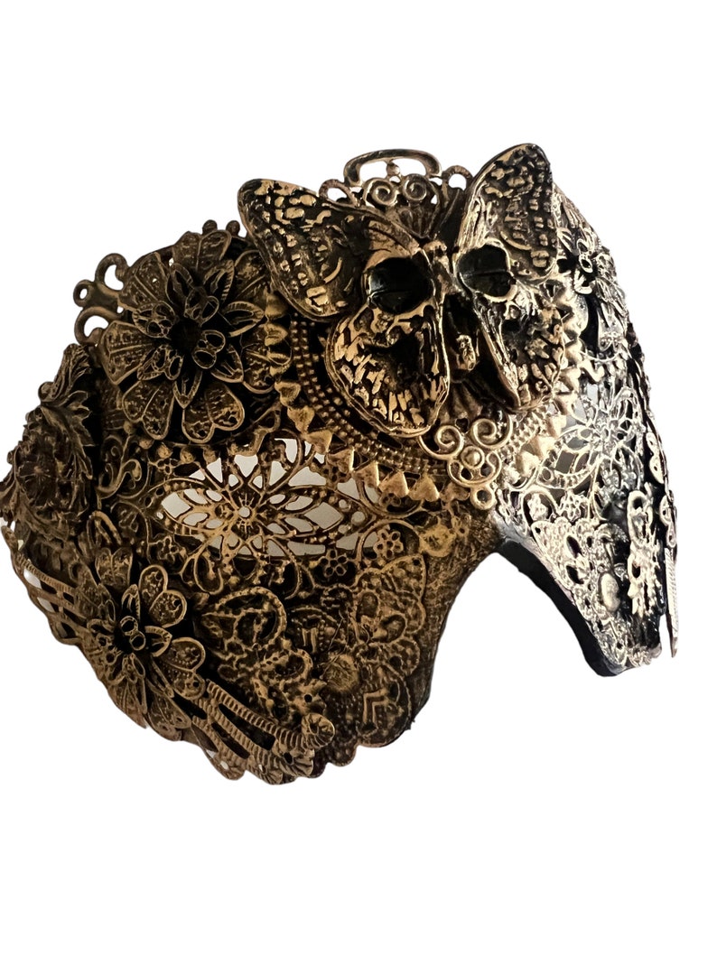 Puede incluir: Una m&aacute;scara de metal dorado con un dise&ntilde;o de calavera y mariposa. La m&aacute;scara est&aacute; intrincadamente detallada y tiene un delicado patr&oacute;n de encaje.