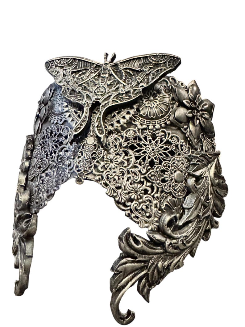 May include: A silver-colored headpiece with intricate floral and butterfly designs. The headpiece features detailed filigree patterns, with a prominent butterfly at the top and floral accents along the sides. The metalwork has a vintage, antique appearance.
