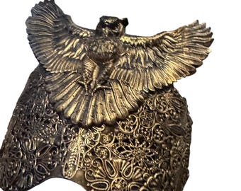 May include: A bronze-colored decorative mask featuring an owl with outstretched wings. The mask has intricate floral and scrollwork patterns. The owl is centered above the mask, with detailed feathering and a textured surface.