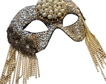 May include: An ornate masquerade mask with a black base, heavily embellished with pearls, rhinestones, and gold-toned accents. The mask features dangling pearl and rhinestone fringe, and a floral embellishment on the side. The overall design is glamorous and elegant.