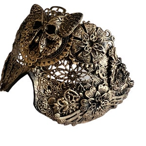 Puede incluir: Una m&aacute;scara de metal dorado con intrincados dise&ntilde;os florales y de mariposas. La m&aacute;scara est&aacute; hecha de metal delicado y calado y tiene una mariposa con forma de calavera en la parte superior.