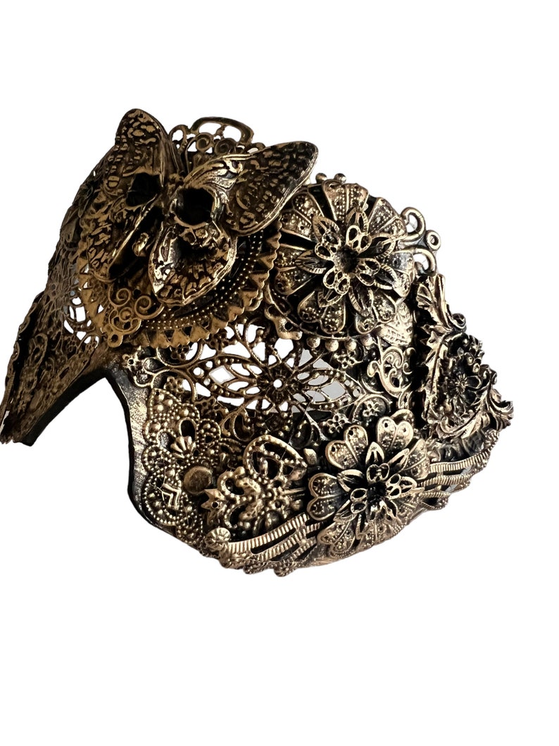 Puede incluir: Una m&aacute;scara de metal dorado con intrincados dise&ntilde;os florales y de mariposas. La m&aacute;scara est&aacute; hecha de metal delicado y calado y tiene una mariposa con forma de calavera en la parte superior.
